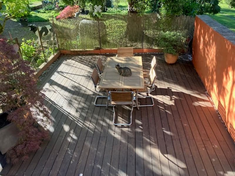 Wooden deck patio with a table, chairs, and a cat sitting on the table. The area is surrounded by a wooden fence, with greenery and a cow visible in the background.