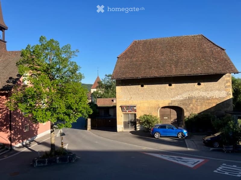 A historic stone building with a traditional roof, surrounded by greenery and a parking area.