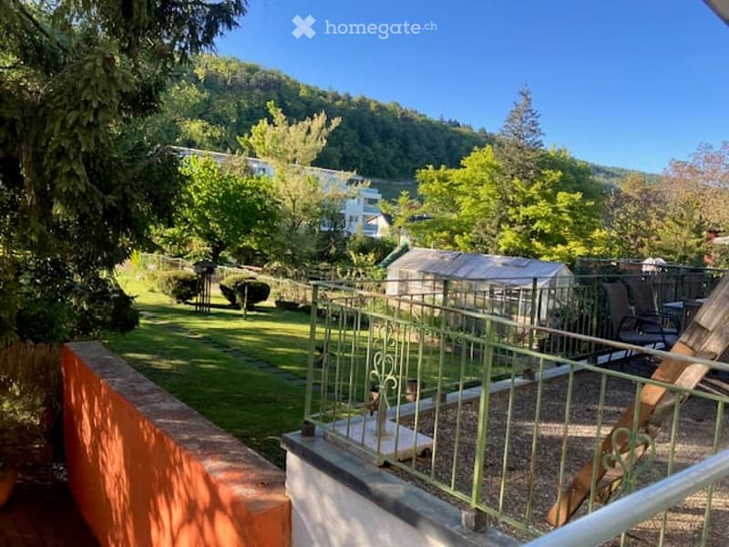 Balcony overlooking a lush garden with trees, a small shed, and distant buildings visible in the background.