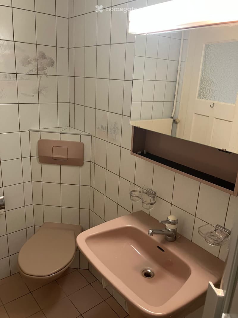 Bathroom with beige tiles, a toilet, a sink with a faucet, a mirror with a built-in shelf, and a cup holder.