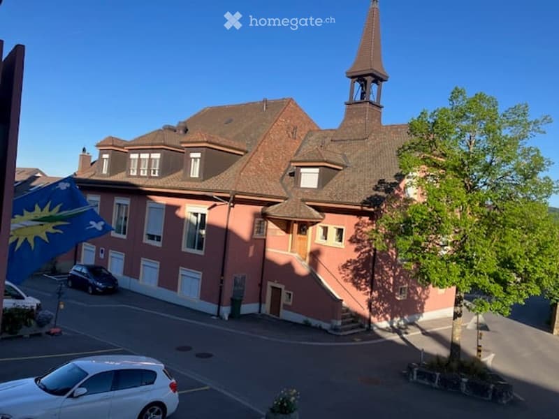 A multi-story building with a red brick facade, multiple windows, and a prominent tower with a spire. The building is surrounded by a paved area with parked cars and a tree.