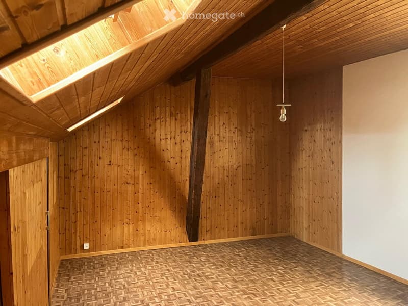 An empty room with wooden walls, a slanted wooden ceiling, and a patterned wooden floor. A single hanging light fixture is visible, and there is a door on one side.