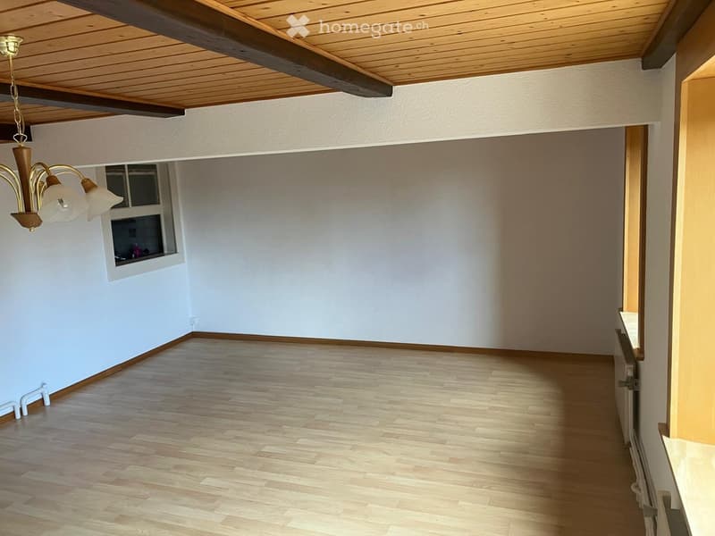 Empty room with wooden flooring, white walls, and a chandelier hanging from the ceiling. There is a window on one wall and a doorway leading to another room.