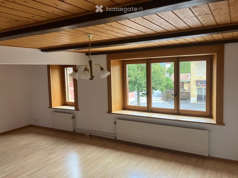 Empty room with wooden flooring, exposed wooden ceiling beams, large windows, radiators, and a hanging light fixture.