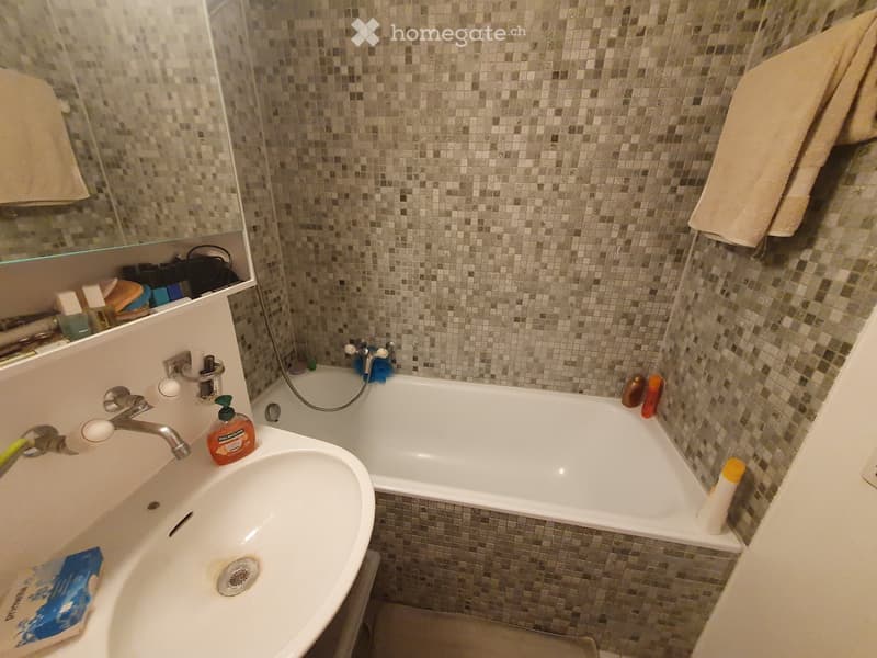 Bathroom with a white sink, bathtub, and mosaic tile walls. A mirror above the sink, and towels hanging on the side.