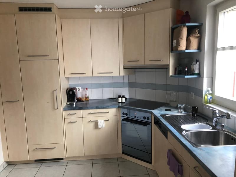 Modern kitchen with light wood cabinets, stainless steel sink, oven, and various appliances on the counter.