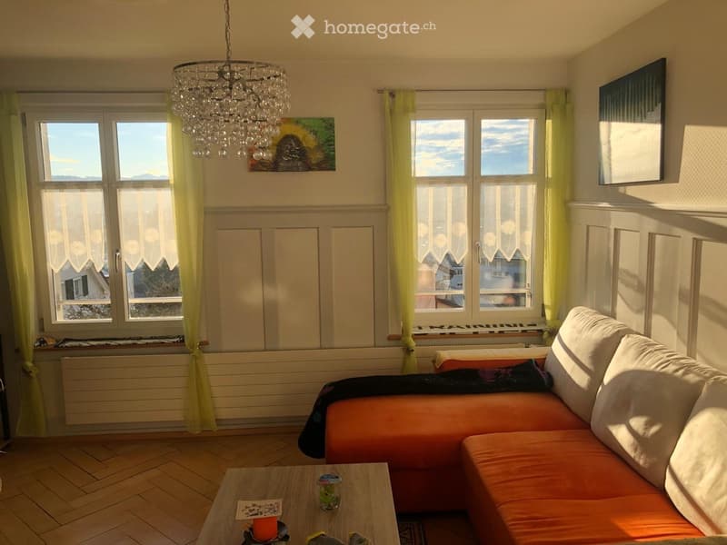 Bright living room with large windows, orange sectional sofa, wooden coffee table, chandelier, and artwork on the walls.