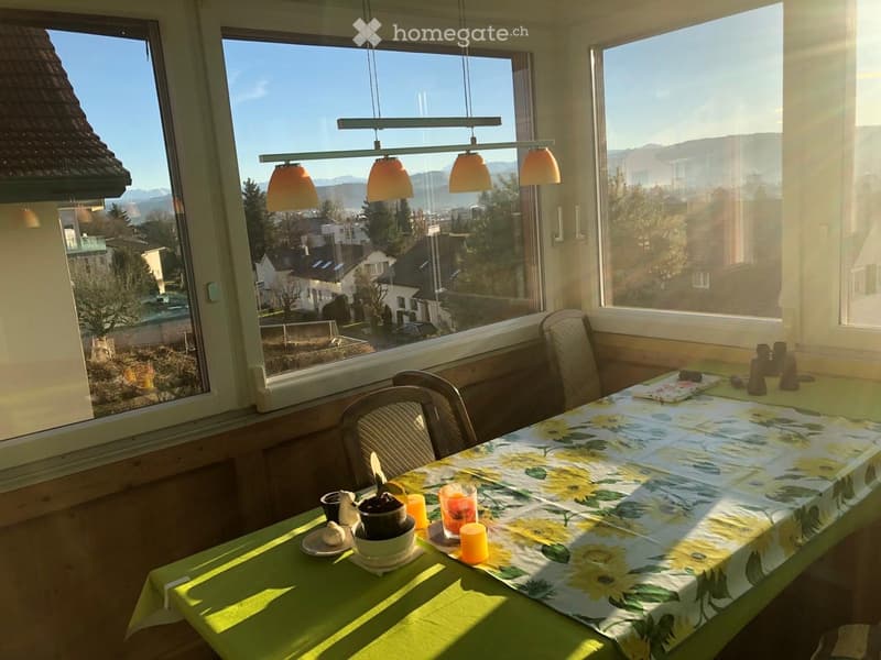 Bright kitchen with large windows overlooking a scenic view, featuring a dining table with a floral tablecloth, chairs, and various decorative items.