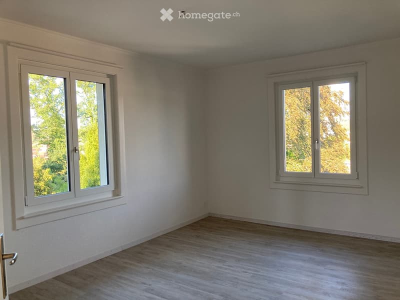Empty room with light wooden flooring, white walls, and two large windows with white frames, offering a view of greenery outside.