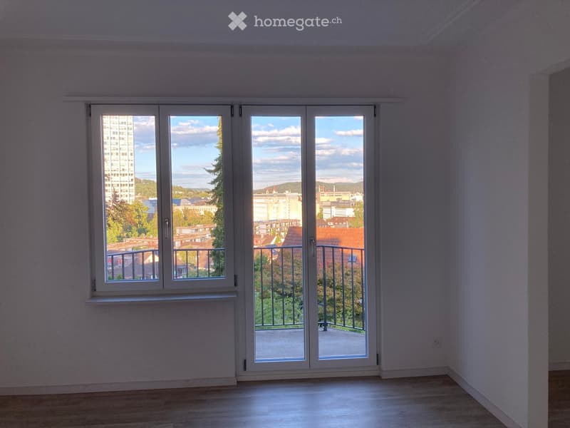 Empty room with large windows and sliding doors leading to a balcony, showing a cityscape view.