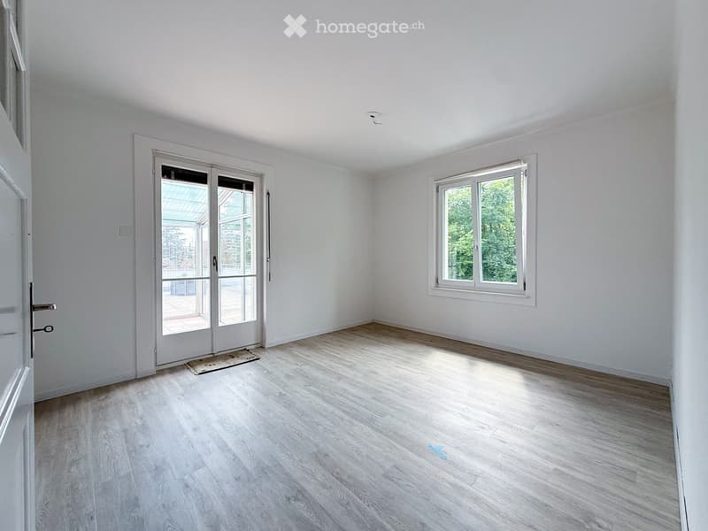 Empty room with white walls, light wooden flooring, a large window, and a sliding glass door leading outside.