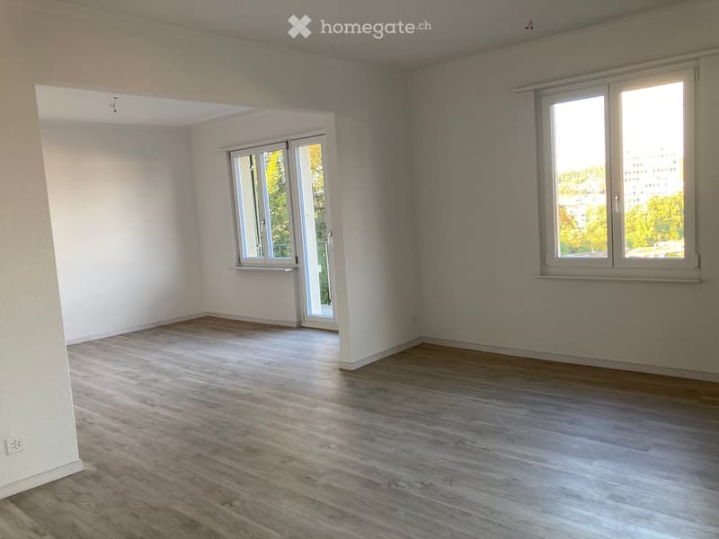 Empty room with wooden flooring, white walls, and large windows with views of trees and buildings.