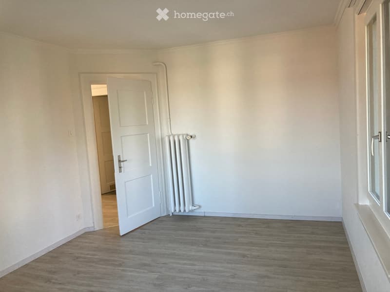 Empty room with light-colored walls, wooden flooring, and a radiator near the door. The door is open, revealing a glimpse of another room with similar flooring.