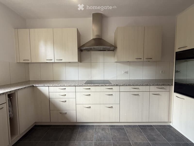 Modern kitchen with light wood cabinets, stainless steel range hood, granite countertop, and dark tiled floor.