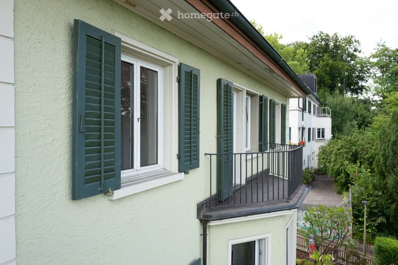 A two-story building with light green exterior walls, dark green shutters, and a small balcony with a black metal railing. The building features multiple windows and a sloped roof with a visible gutter.