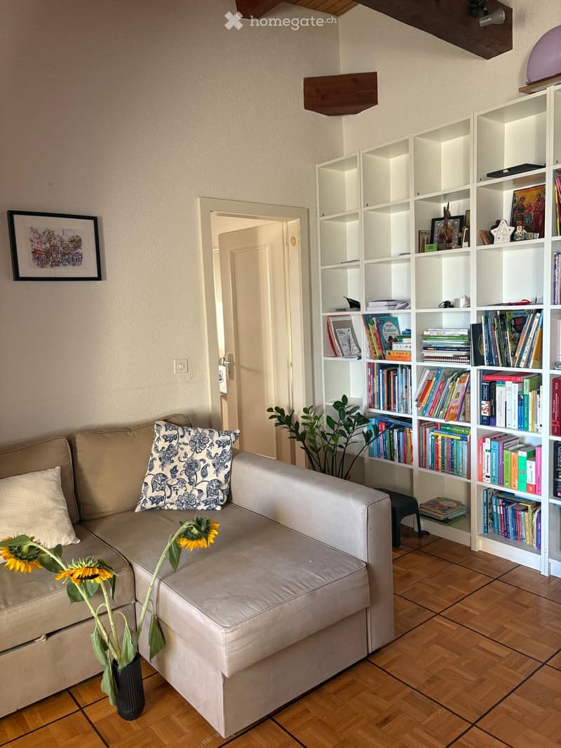 Living room with a beige sectional sofa, decorative pillows, a bookshelf filled with books and decorative items, a small black stool, a potted plant, and a vase with sunflowers on the floor.