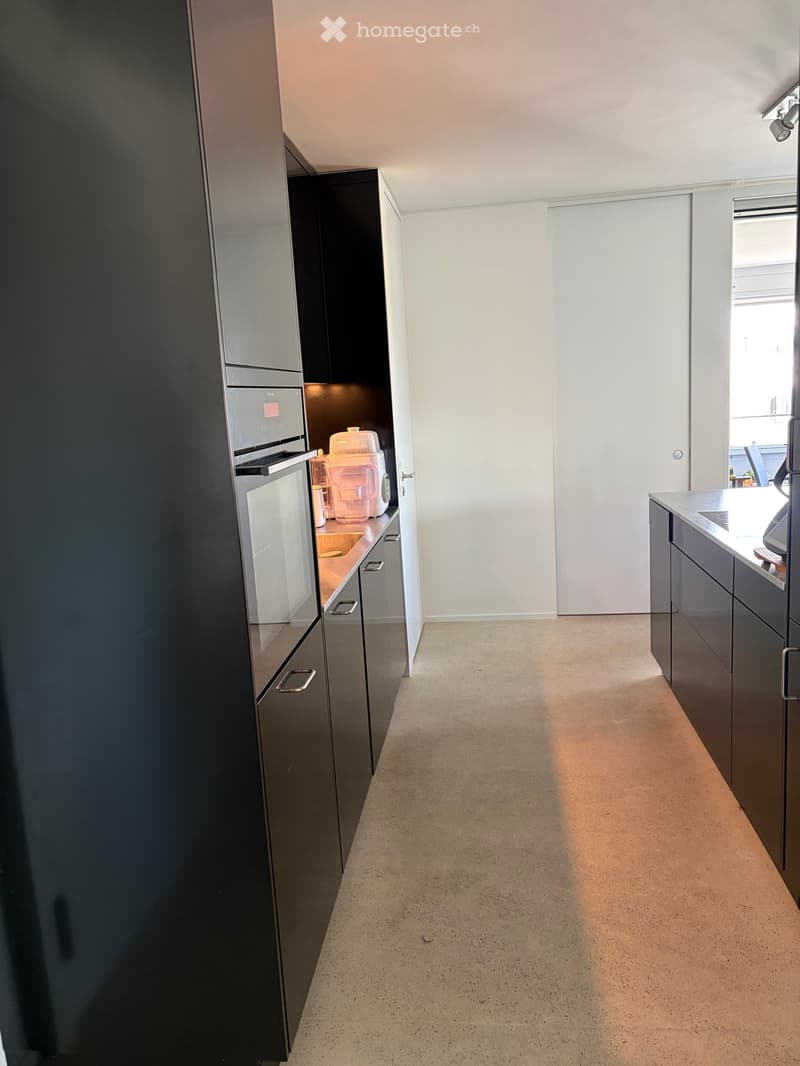 Modern kitchen with dark grey cabinets, light-colored floor, and built-in appliances including a refrigerator, microwave, and oven. The space is clean and well-lit with natural light coming from a window on the right.