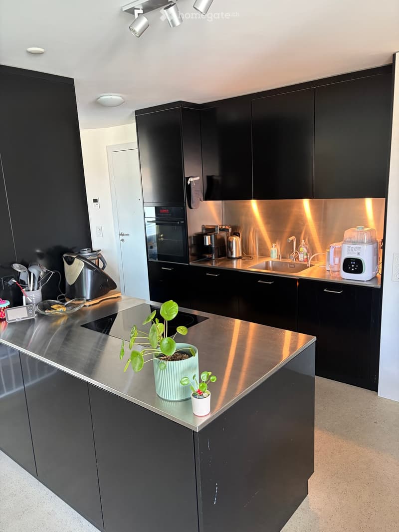 Modern kitchen with black cabinets, stainless steel countertops, and various appliances including an oven, coffee maker, toaster, and blender. There is a small plant on the island.