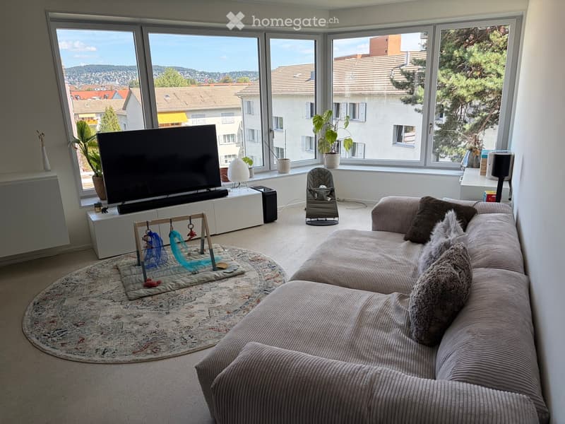 Bright living room with large windows offering a city view, featuring a sectional sofa, television, side table, floor lamp, and decorative plants.