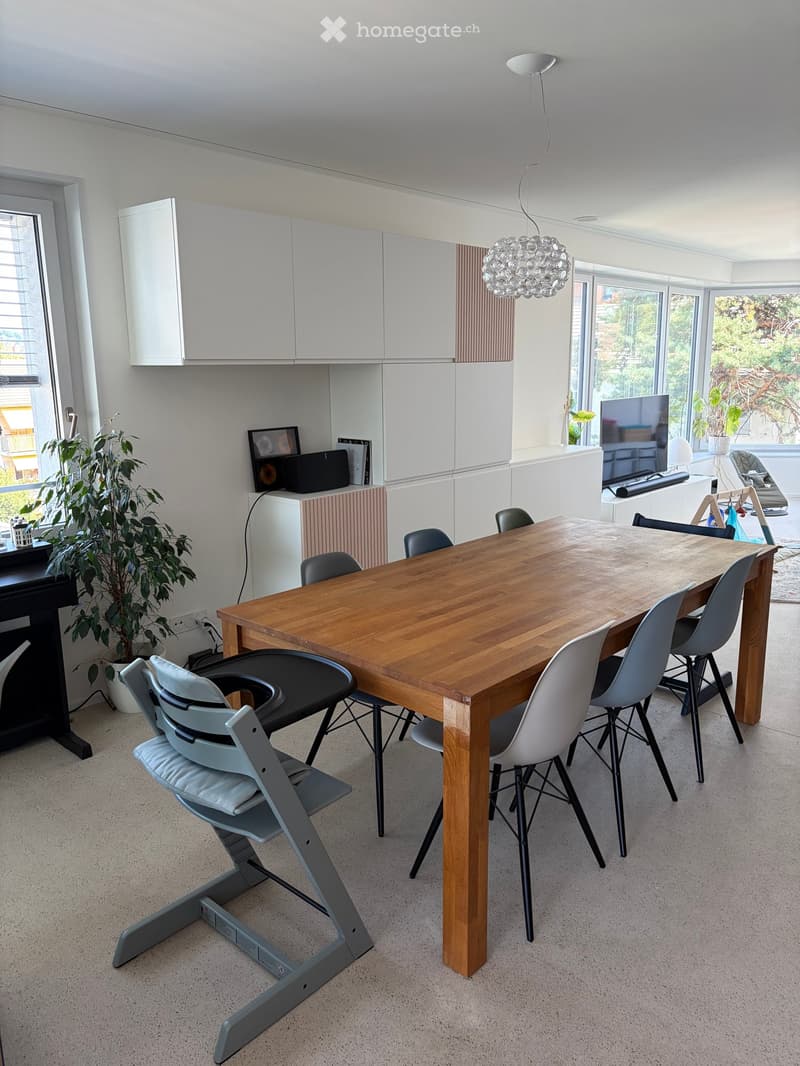 Modern dining area with a wooden dining table, multiple chairs including a baby chair, white cabinets, a large window with blinds, and a decorative lamp.