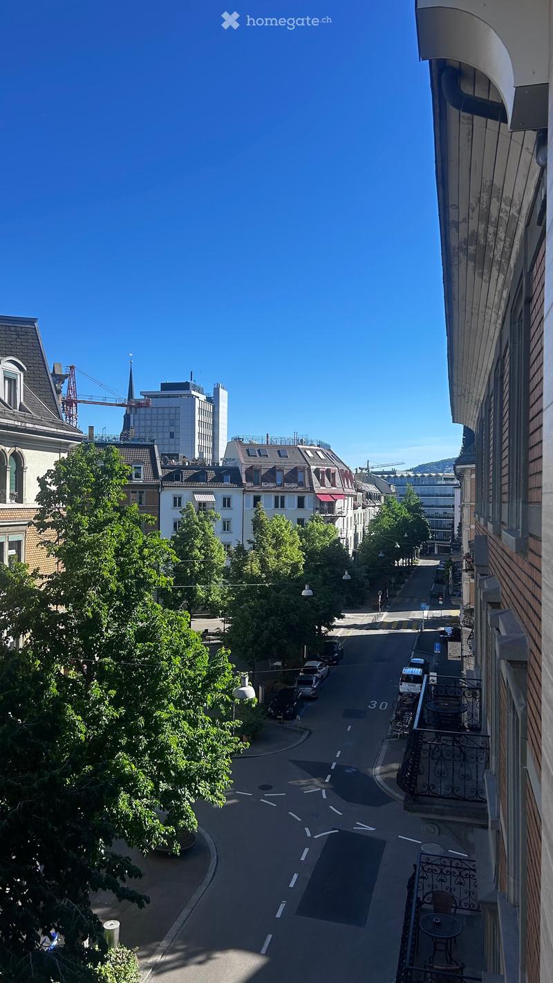 Balcony overlooking a quiet urban street lined with trees and parked cars, with buildings and a distant spire visible in the background.