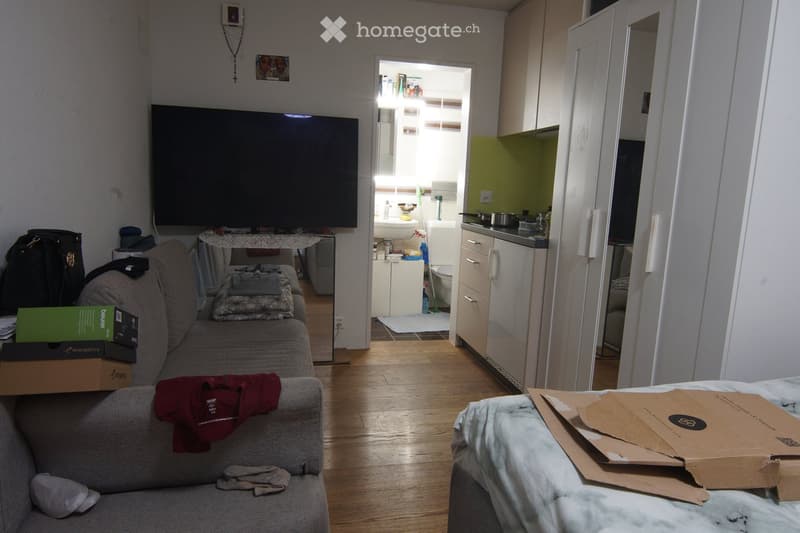 A furnished living room with a sofa, a small table, and a television. A kitchen area is visible in the background with a sink, refrigerator, and cabinets. There are boxes and some clothing items on the sofa and a bed.