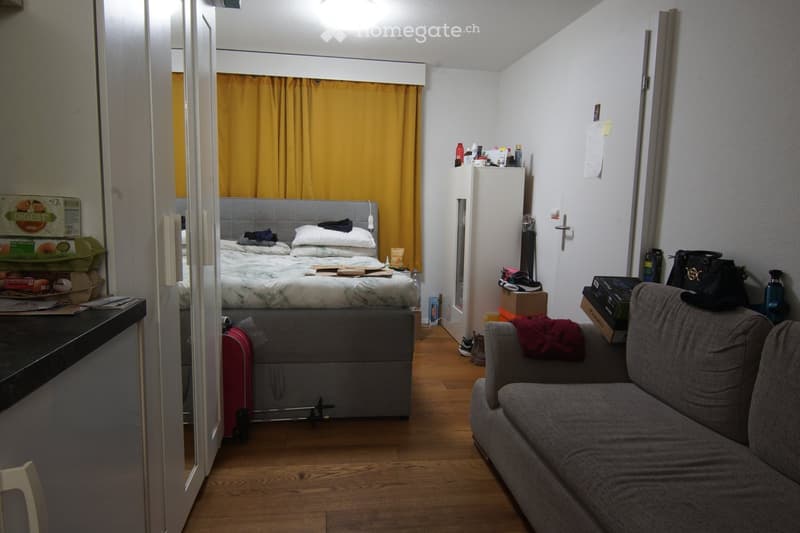 A small, furnished room with a bed, sofa, and a refrigerator. The room features wooden flooring, a grey sofa with a red blanket, a bed with pillows and a duvet, and various personal items including a suitcase, handbag, and laptop.
