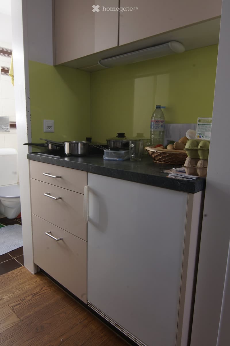 Modern kitchen with light-colored cabinets, black countertop, and various kitchen items including a pot, frying pan, cup, bottle, wicker basket, and eggs on the counter.