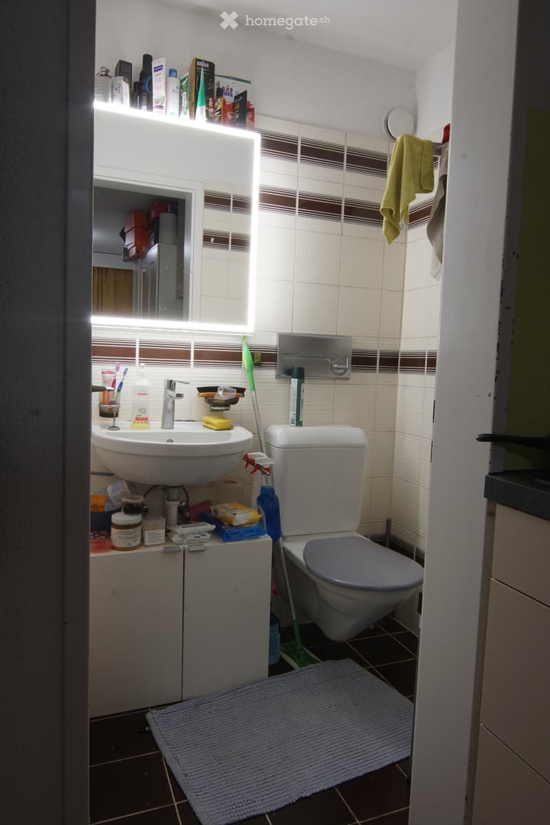A small bathroom with a white sink, silver faucet, toilet, and a cabinet below the sink. The walls are tiled with brown and white tiles, and there is a towel hanging on the side. Cleaning supplies and personal care items are visible on the sink and cabine