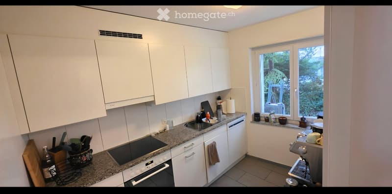 Modern kitchen with white cabinets, granite countertops, and stainless steel appliances. Features a sink, oven, dishwasher, coffee machine, and various kitchen items on the counter.