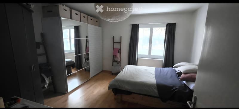 A well-lit bedroom with a wooden floor, a large bed with pillows and blankets, a mirrored wardrobe, and a window with dark curtains.