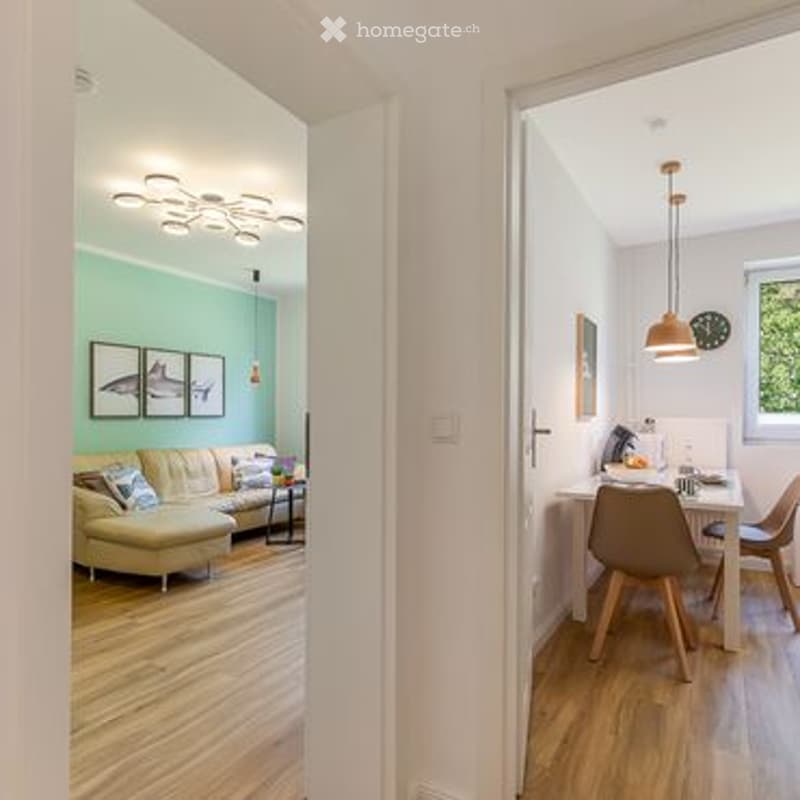 Open-plan living and dining area with light green walls, wooden flooring, a cream-colored sofa, a coffee table, a dining table with chairs, and pendant lights.