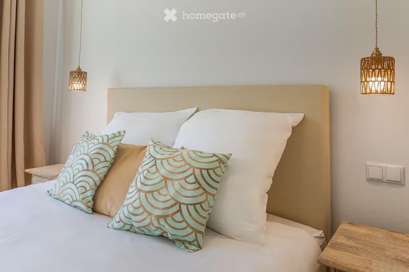 A modern bedroom with a large bed, decorative pillows, and two hanging pendant lights. The room features a beige headboard and light-colored walls.