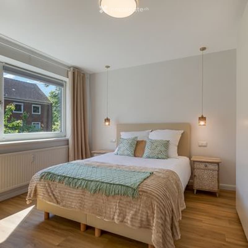 Bright bedroom with wooden flooring, large window with view, bed with pillows and blanket, nightstand, and hanging pendant lights.