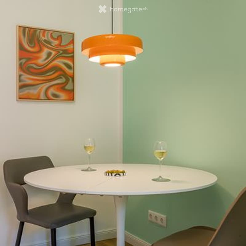 A small, modern dining area with a white round table, two chairs, two wine glasses, a small toy car, and an abstract picture on the wall. An orange pendant light hangs from the ceiling.
