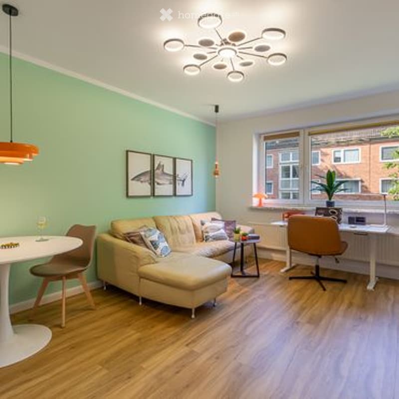 Bright living room with light green walls, wooden flooring, and large windows. Features a cream sectional sofa, coffee table, dining table with chairs, pendant light, picture frames, and a small desk with a chair.