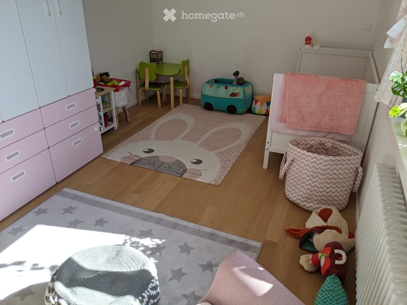 A well-lit children's room with a wooden floor, two rugs, a wardrobe, a small table with chairs, a toy car, and various toys scattered around.
