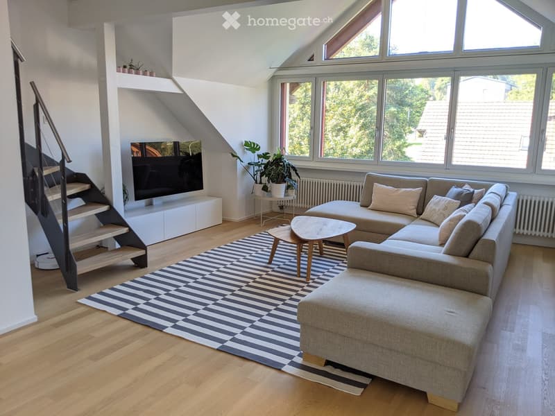 Bright living room with wooden flooring, large windows, a sectional sofa, coffee table, TV, and a staircase leading upwards.