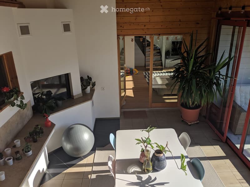 A modern, well-lit room with a large window, a white table with chairs, a large exercise ball, and several potted plants. The room has a tiled floor and a sliding glass door leading to another area.