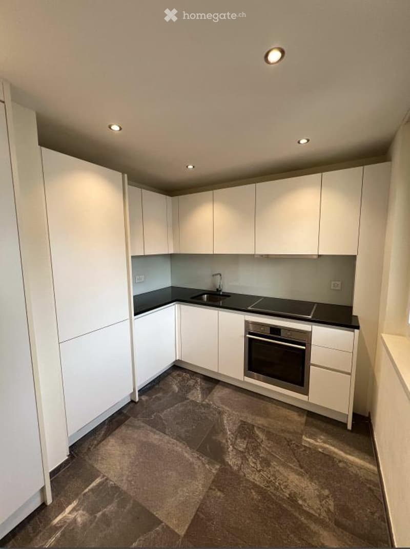 Modern kitchen with white cabinets, black countertops, and built-in appliances including a sink, faucet, and oven. The floor is tiled in a dark, textured pattern.