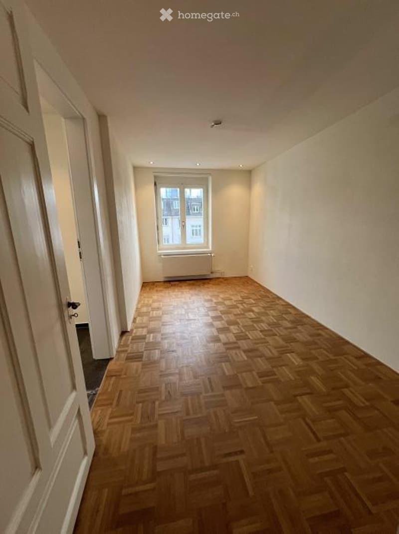 Empty room with wooden parquet flooring, white walls, and a single window with a radiator below it. The room has a door on the left side and recessed ceiling lights.