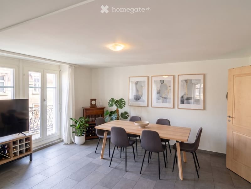 Modern dining room with a wooden dining table, six chairs, a wine rack, a television, and decorative plants and artwork.