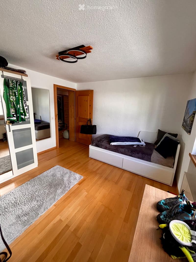 A modern bedroom with a wooden floor, a white bed with dark bedding, a grey rug, a handbag on the floor, a mirror, and a wardrobe visible in the background.