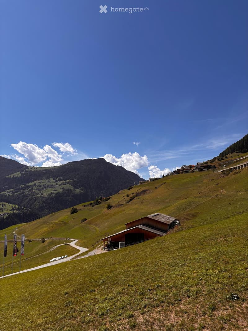 A scenic mountain landscape with a wooden barn on a grassy hillside, surrounded by rolling green fields and distant peaks under a clear blue sky.