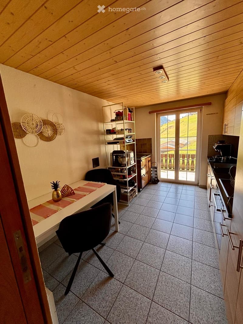 Bright, open-plan room with a wooden ceiling, tiled floor, and large sliding glass door leading to a balcony with a scenic view. Includes a dining table with chairs, a shelf with kitchen appliances, and a modern kitchen area with cabinets and a sink.