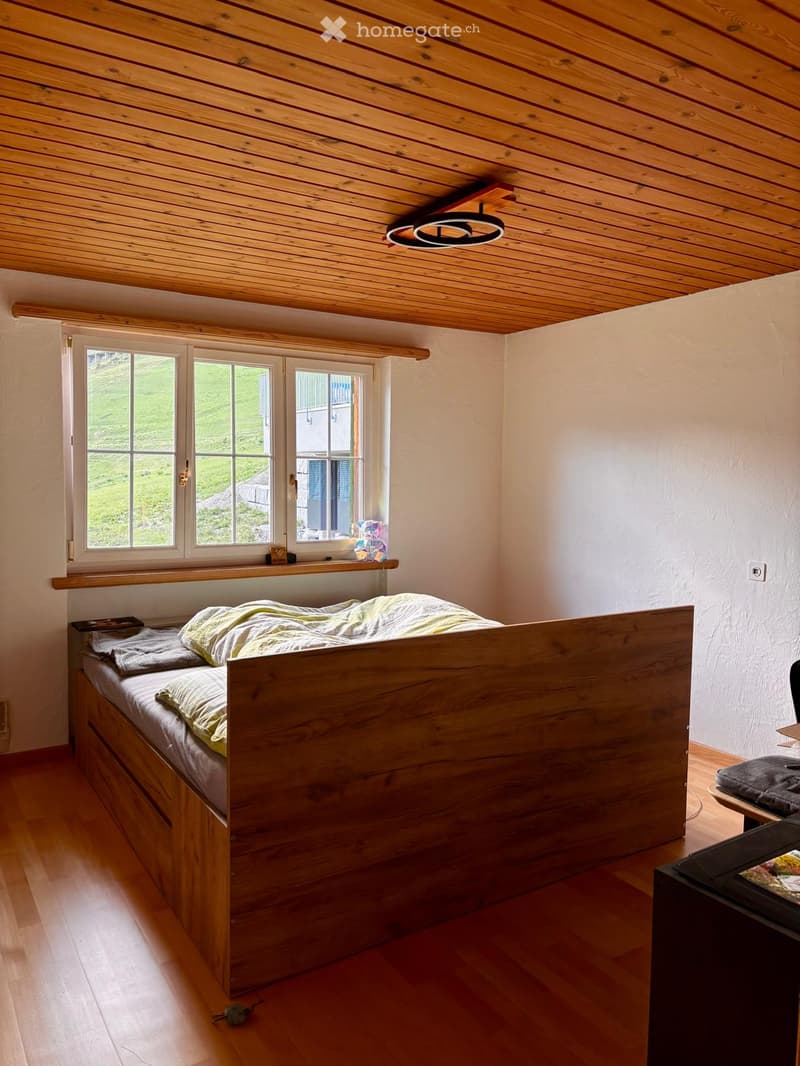 Cozy bedroom with wooden bed, nightstand, and shelf. Large windows offer a view of the green landscape.