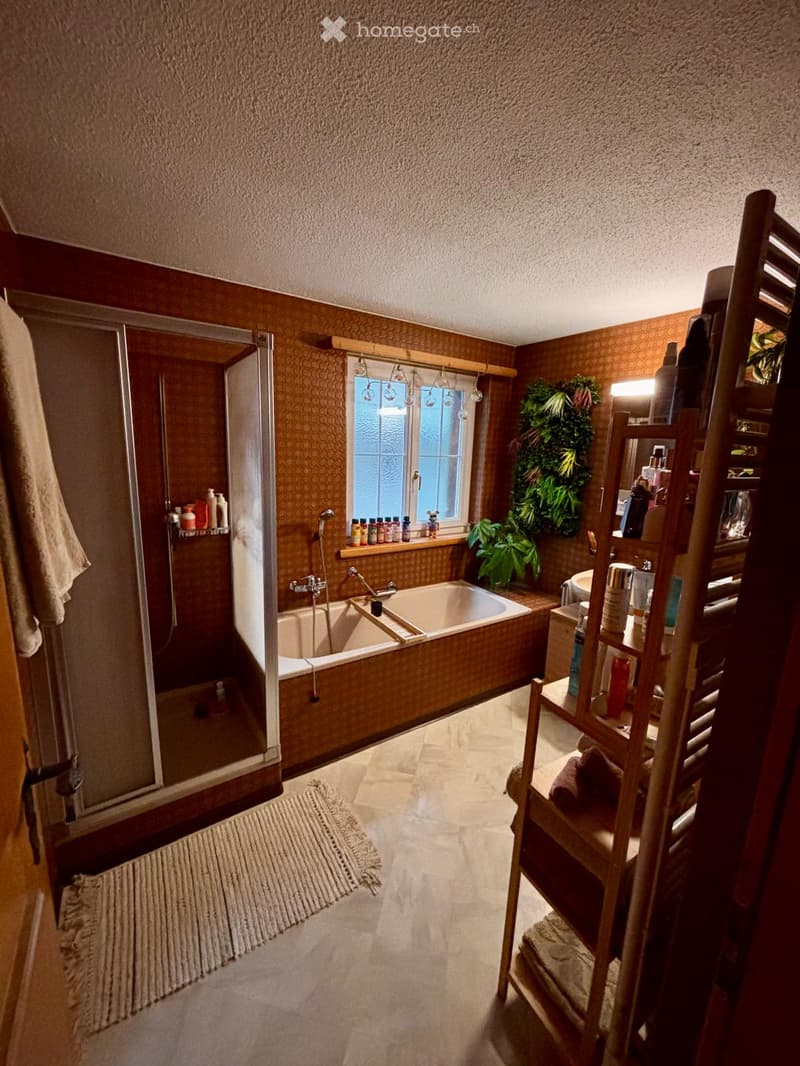 Bathroom with a bathtub, shower, sink, mirror, and shelves holding toiletries. Features a window with a view, a small plant, and a towel on the floor.