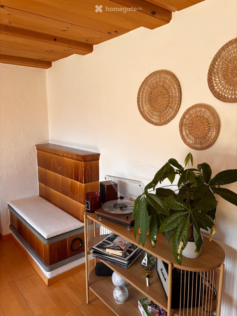 A cozy, well-furnished room with a wooden bed, a round wooden table holding a large leafy plant, a record player, and decorative wall hangings.
