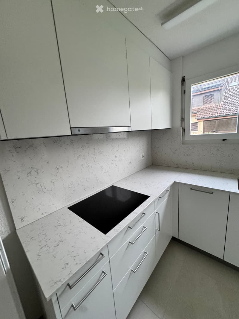 Modern kitchen with white cabinets, marble countertop, and built-in induction stove. Natural light from a window on the right.