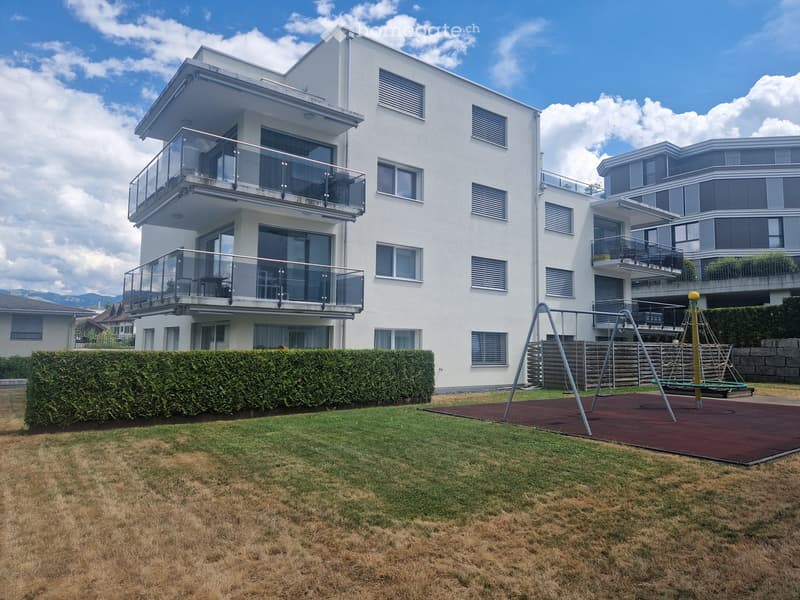 Modern two-story white building with large windows and balconies, surrounded by a well-maintained lawn and a playground area with swings and a climbing frame.